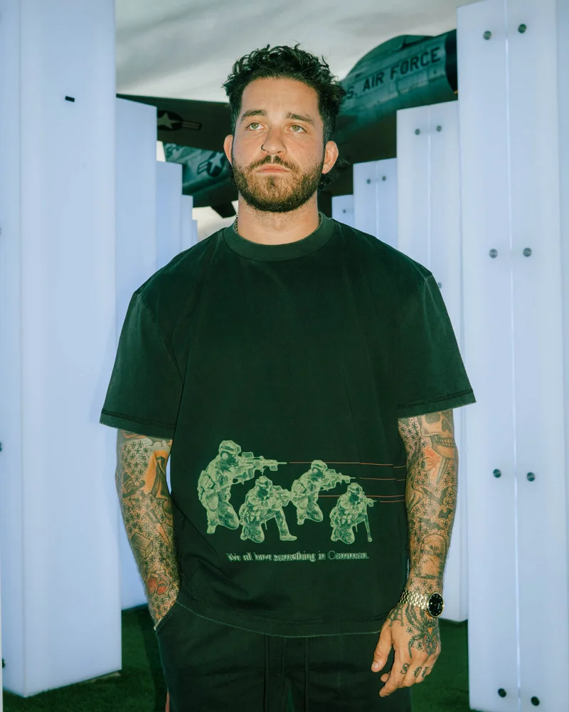 A tattooed model in a green Commons graphic tee standing in a white studio set.