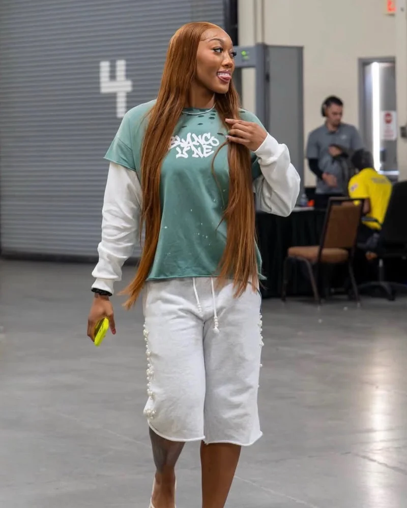 A model in a green Commons graphic tee and white shorts walking through an arena hallway.