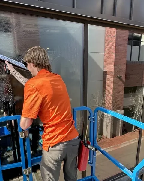 Cleaner in an orange shirt washing exterior windows from a blue elevated work platform.